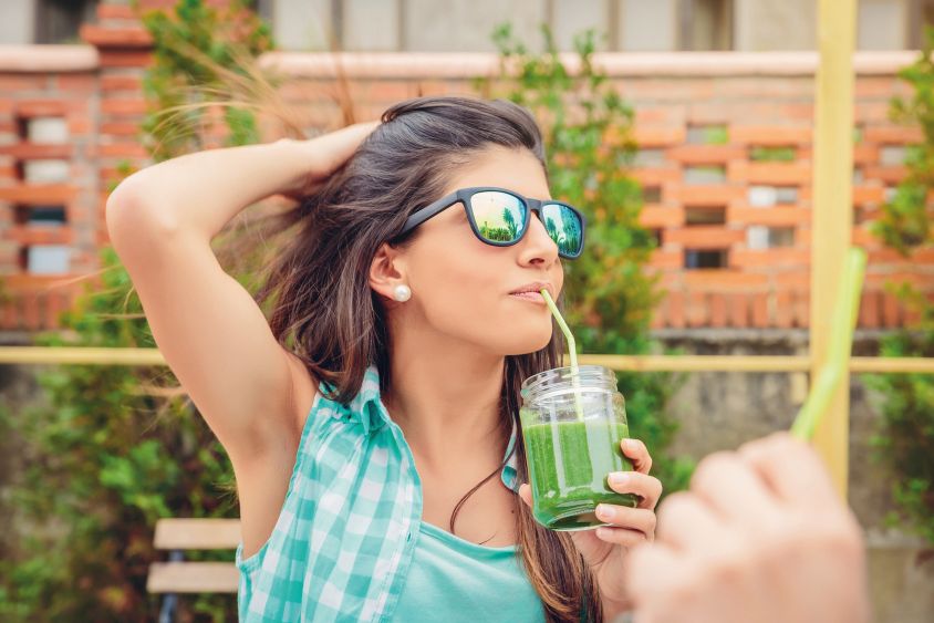 Beautiful young woman with sunglasses drinking green vegetable smoothie with straw in a summer day outdoors. Healthy organic drinks concept.